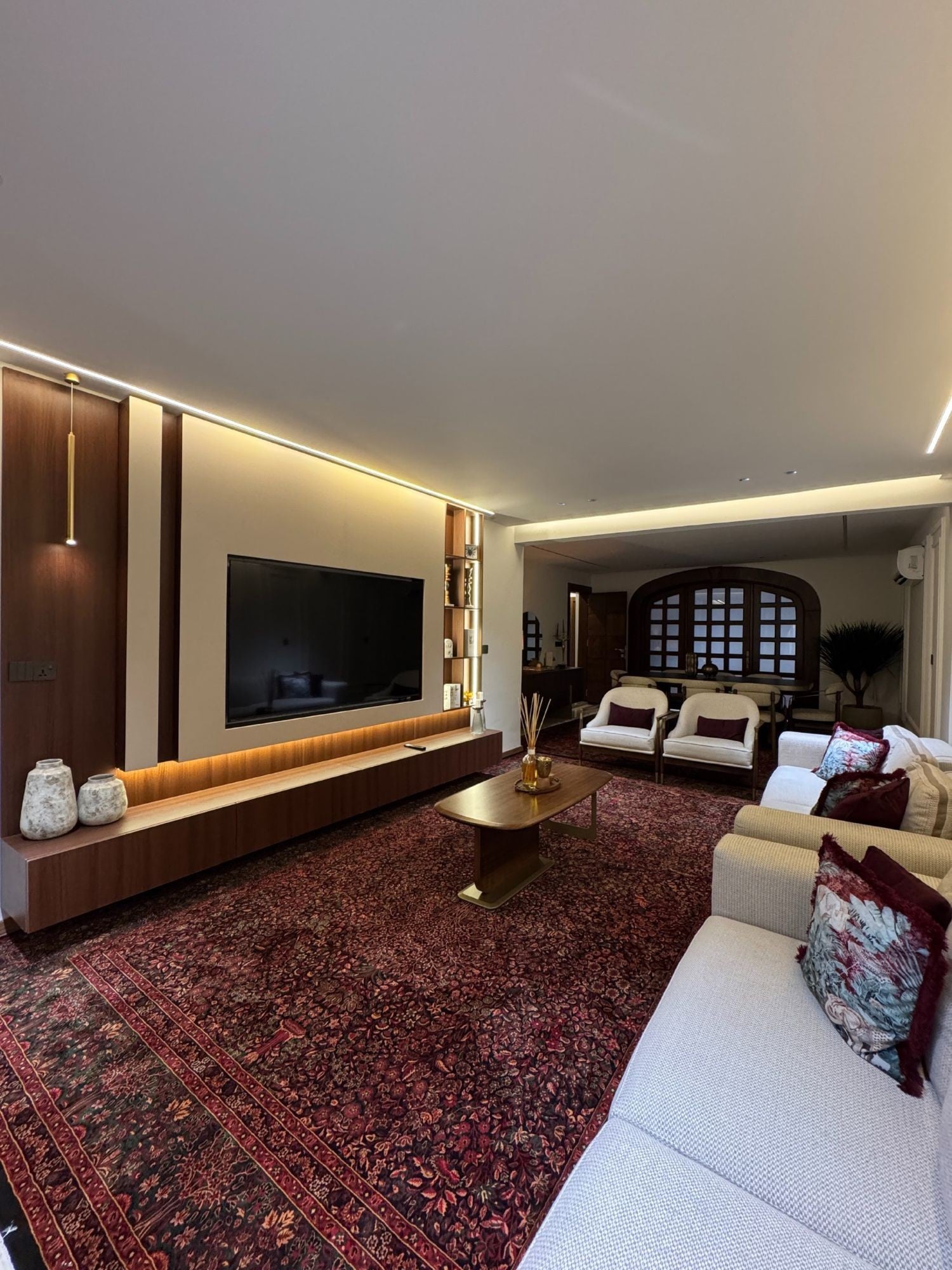 صالة بجدار شاشة من خشب MDF بلون الجوز وإطار بيج ورفوف مضاءة وجلسات بيضاء وسجادة